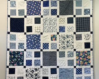 May include: A square quilt with a patchwork design, featuring various blue floral and patterned fabrics. The quilt is framed with a dark blue border and white squares. It is hung on a wooden rod with white fabric ties.