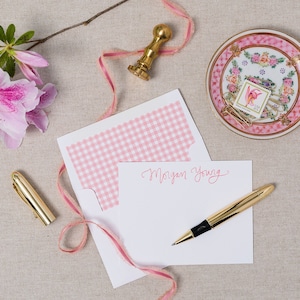 May include: A white envelope with a pink and white gingham pattern on the flap. The envelope is open and a white card with the name "Morgan Young" written in pink ink is visible. A gold pen and a gold stamp are also in the image.