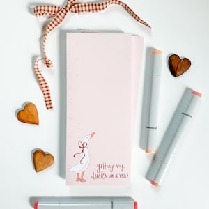 May include: A pink notebook with a goose illustration and the phrase "getting my ducks in a row." The notebook is surrounded by gray markers, wooden heart shapes, and a brown and white checkered ribbon.