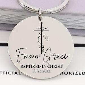 Könnte beinhalten: Ein silberfarbener runder Schlüsselanhänger mit Kreuz- und Blumendesign. Der Schlüsselanhänger trägt den Namen "Emma Grace" und den Text "BAPTIZED IN CHRIST 03.25.2022" in einer Schreibschrift. Der Schlüsselanhänger ist an einem silbernen Ring befestigt.