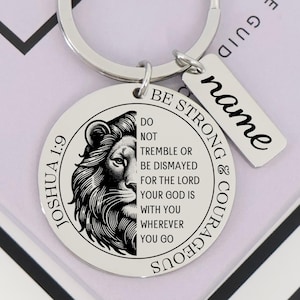 Puede incluir: Un llavero plateado con un colgante redondo que presenta una cabeza de león detallada y el texto "Joshua 1:9". El llavero también incluye una pequeña etiqueta rectangular con la palabra "name" y un anillo. El texto "Be Strong & Courageous" también está en el colgante.