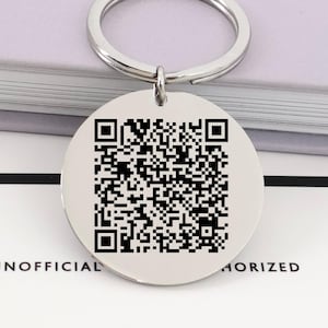 May include: A silver keyring with a circular pendant displaying a black and white QR code. The keyring has a silver loop for attaching keys. The background includes a book and text that reads "UNOFFICIAL".