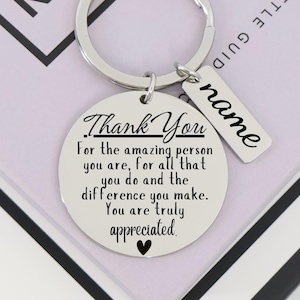 May include: A silver-colored keychain with a round pendant and a rectangular tag. The round pendant has the words "Thank You" and a message of appreciation. The rectangular tag has the word "name".