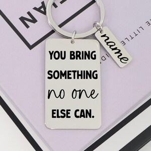 Puede incluir: Un llavero rectangular plateado con el texto negro "YOU BRING SOMETHING no one ELSE CAN.". Una etiqueta más pequeña con la palabra "name" está adjunta. El llavero tiene un anillo circular.