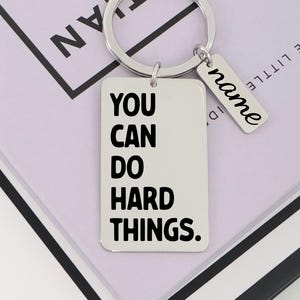 Puede incluir: Un llavero rectangular plateado con las palabras "YOU CAN DO HARD THINGS" en negro. Una etiqueta más pequeña con la palabra "name" está adjunta. El llavero está sobre una superficie de color púrpura claro.