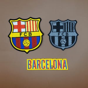 May include: Two embroidered patches featuring the FC Barcelona crest, one in vibrant colors and the other in grayscale. Below, a rectangular patch spells out "BARCELONA" in yellow with blue and red accents. The patches are on a brown surface.