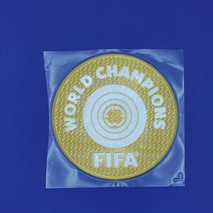 May include: A gold-colored patch with the words "WORLD CHAMPIONS" and "FIFA" in white lettering. The patch is circular with a concentric circle design in the center. It is sealed in a clear plastic bag.