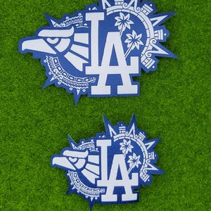 May include: Two blue and white patches featuring the letters "LA" in a stylized font. The patches have a circular design with an eagle head, palm trees, and geometric patterns. The patches are on a green background.