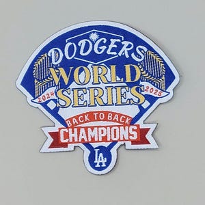 May include: A patch featuring the text "Dodgers World Series" in gold lettering on a blue background. The patch also includes the text "Back to Back Champions" in red and white, with the years 2024 and 2025.