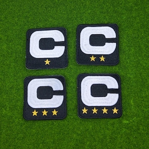 May include: Four black embroidered patches with a white "C" design and gold stars. The patches are square-shaped and arranged on a green surface. Each patch has a different number of gold stars.