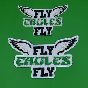 May include: Two embroidered patches with the text "FLY EAGLES FLY" in black and green. The patches feature white wings with black and tan detailing. The background is a solid green color.