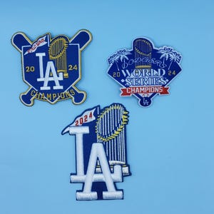 May include: Three embroidered patches featuring the Los Angeles Dodgers logo and the text "World Series Champions." The patches are blue, white, and gold, with the year 2024 displayed prominently. The patches are on a light blue background.