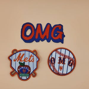 May include: Three embroidered patches with baseball themes. One patch says "OMG" in red letters with a blue outline. Another patch says "Mets" with baseball bats and a "Homerun Queens" logo. The third patch is a baseball with "OMG" and "Mets" text.