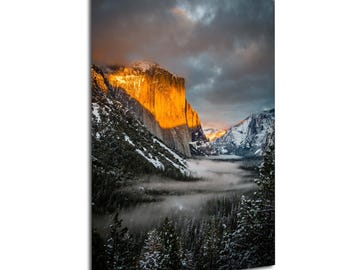 Könnte beinhalten: Landschaftsfotografie eines Gebirges bei Sonnenuntergang. Die Sonne beleuchtet eine große Felswand mit goldenem Schein. Schnee bedeckt die Berggipfel, und der Vordergrund zeigt einen Wald und Nebel.
