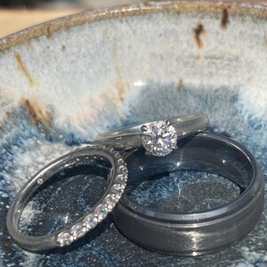 May include: Three rings rest in a small, blue and white ceramic bowl. One ring features a large, clear gemstone, while another is adorned with smaller stones. The third ring is a simple, dark gray band.