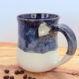 May include: A handmade ceramic mug with a heart-shaped accent on the handle. The mug features a two-tone design, with a white base and a dark blue, glossy glaze on the upper portion. Coffee beans are scattered on the wooden surface.