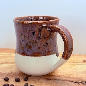 May include: A handmade ceramic mug with a heart-shaped accent on the handle. The mug features a two-tone design, with a white base and a speckled purple and brown upper section. Coffee beans are scattered around the mug.