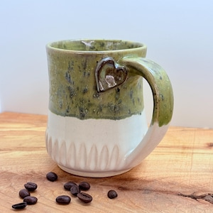 May include: Handmade ceramic mug with a heart-shaped handle attachment. The mug features a two-tone design, with a green speckled top and a white, textured bottom. Coffee beans are scattered on the wooden surface next to the mug.