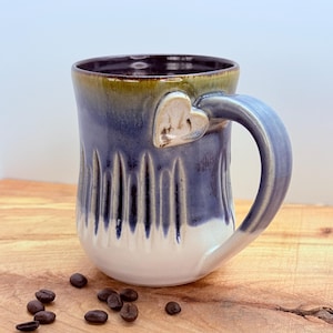 May include: A handmade ceramic mug with a heart-shaped accent on the handle. The mug features a gradient of colors, transitioning from a dark blue to white, with a brown interior. Coffee beans are scattered on the wooden surface.