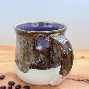 May include: A handmade ceramic coffee mug with a heart-shaped handle. The mug features a dark brown and gold glaze over a white base, with a deep purple interior. Coffee beans are scattered on the wooden surface.