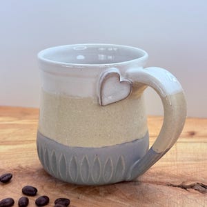 May include: A handmade ceramic mug with a heart-shaped accent on the handle. The mug features a color-blocked design with white, tan, and gray sections. Coffee beans are scattered on the wooden surface next to the mug.