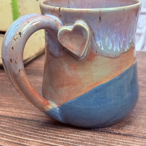 May include: A ceramic mug featuring a multi-colored glaze with blended tones of blue, orange, and lavender. The mug has a curved handle and a small, raised heart-shaped accent attached to the side where the handle meets the body of the mug.