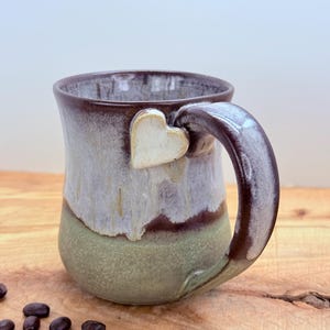 May include: Handmade ceramic mug with a heart-shaped accent. The mug features a brown rim and handle, with a light green and brown body. The heart is a cream color. Coffee beans are scattered on the wooden surface.