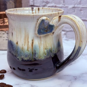 May include: A ceramic mug featuring a two-tone glaze with a cream-colored top that transitions into a deep dark brown base. A small, textured blue heart is attached to the upper side near the handle. Several roasted coffee beans are scattered.