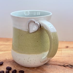 May include: A handmade ceramic mug with a heart-shaped accent on the handle. The mug features a two-tone design with a white top and bottom, and a green center. Coffee beans are scattered on the wooden surface.