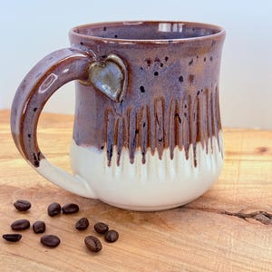 May include: A handmade ceramic mug with a heart-shaped accent on the handle. The mug features a two-tone design with a white base and a speckled purple glaze that drips down. Coffee beans are scattered on the wooden surface.