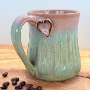 May include: A handmade ceramic mug with a heart-shaped accent on the handle. The mug features a gradient of colors, including green, pink, and brown. Coffee beans are scattered on the wooden surface next to the mug. The mug is designed for hot beverages.
