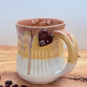 May include: Handmade ceramic mug with a heart-shaped accent on the handle. The mug features a cream, yellow, and brown color scheme with vertical ribbed detailing. Coffee beans are scattered on the wooden surface near the mug.