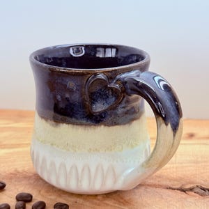 May include: A handmade ceramic mug with a heart-shaped handle. The mug features a gradient of colors, starting with a dark blue rim, transitioning to a tan middle, and ending with a white, textured base. Coffee beans are scattered on the wooden surface.
