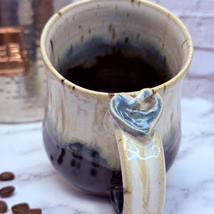 May include: A ceramic mug with a two-tone glaze featuring a cream-colored upper section and a dark brown lower section. The handle is accented with a sculpted heart shape at the top, and the interior is dark, showing a smooth, glazed finish.