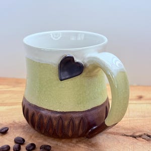 May include: A handmade ceramic mug with a heart-shaped accent on the handle. The mug features a color scheme of white, light green, and brown. The base has a textured design. Coffee beans are scattered on the wooden surface.