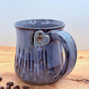 May include: A handmade ceramic mug with a blue and brown glaze. The mug features a heart-shaped accent on the handle and vertical textured lines on the body. Coffee beans are scattered on the wooden surface.