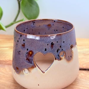 May include: A ceramic candle holder with a heart-shaped cutout. The holder has a speckled purple and brown glaze on the top half, transitioning to a cream-colored base. Small holes are around the heart.