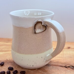 May include: A handmade ceramic mug with a heart-shaped accent on the handle. The mug features a two-tone design with a white rim and handle, a blush pink middle section, and a speckled cream base. Coffee beans are scattered on the wooden surface.