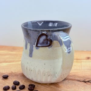 May include: A handmade ceramic mug with a heart-shaped accent on the handle. The mug features a speckled cream and gray glaze with a dark gray rim. Coffee beans are scattered on the wooden surface near the mug.