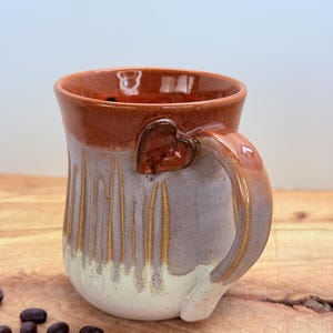 May include: A handmade ceramic mug with a heart-shaped handle. The mug features a two-tone design with a brown rim and handle, and a gray and white body with vertical lines. Coffee beans are scattered on the wooden surface.