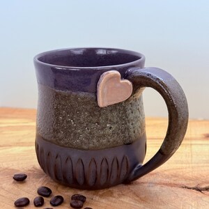 May include: A handmade ceramic mug with a heart-shaped accent on the handle. The mug features a dark purple rim, a textured olive green middle section, and a dark brown base. Coffee beans are scattered on the wooden surface.