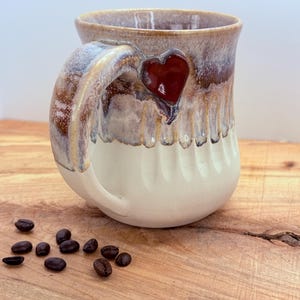 May include: A handmade ceramic coffee mug with a heart-shaped embellishment. The mug features a cream-colored base, a brown and tan glazed top, and a textured handle. Dark coffee beans are scattered on the wooden surface.