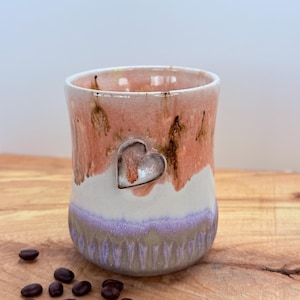 May include: A handmade ceramic mug with a heart-shaped accent. The mug features a color palette of peach, white, and lavender, with brown accents. Coffee beans are scattered on the wooden surface. The mug is approximately 3.5 inches tall.