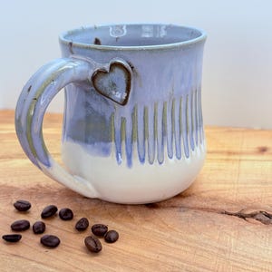 May include: A handmade ceramic mug with a light blue glaze and a white base. The mug features a heart-shaped accent and vertical grooves. Coffee beans are scattered on the wooden surface next to the mug.