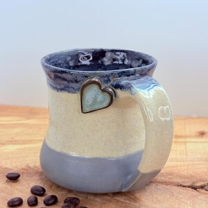 May include: A handmade ceramic mug with a heart-shaped accent. The mug features a blue rim, a cream-colored middle section, and a gray base. The handle is cream-colored. Coffee beans are scattered on the wooden surface.