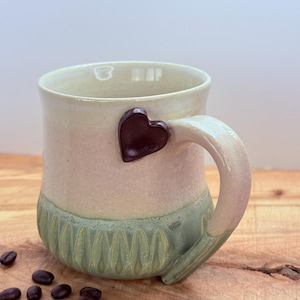 May include: A handmade ceramic mug with a heart-shaped accent. The mug features a light pink upper section, a green textured base, and a dark brown heart. The handle is a light pink color. Coffee beans are scattered on the wooden surface.