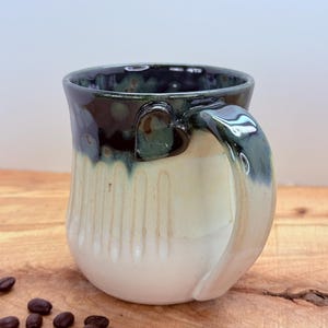 May include: A handmade ceramic mug with a heart-shaped handle. The mug features a white base that fades to a light beige, with a dark teal rim and handle. Coffee beans are scattered on the wooden surface.