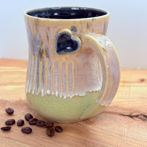 May include: A handmade ceramic mug with a heart-shaped cutout on the side. The mug features a ribbed design, a green base, and a tan and brown glaze. Coffee beans are scattered on the wooden surface.