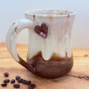 May include: A handmade ceramic mug with a heart-shaped accent. The mug features a white and brown glaze, with a heart-shaped embellishment on the side. Coffee beans are scattered on the wooden surface. The mug is approximately 4 inches tall.
