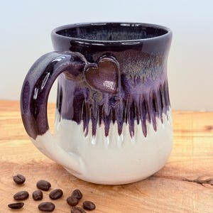 May include: A handmade ceramic coffee mug with a heart-shaped accent on the handle. The mug features a two-tone design with a dark purple top that drips down to a white base. Coffee beans are scattered on the wooden surface.
